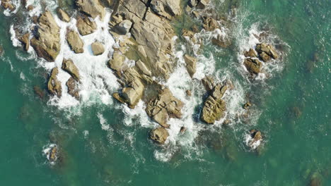 Drone-top-down-view-of-sea-waves-hitting-a-rocky-coast,-Praia-Armacao,-Florianopolis,-Santa-Catarina,-Brazil