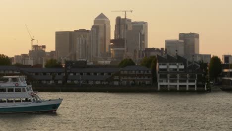 El-Distrito-Financiero-Canary-Wharf-De-Londres-Con-Un-Barco-De-Recreo-Pasando-Por-El-Río-Támesis-Al-Atardecer