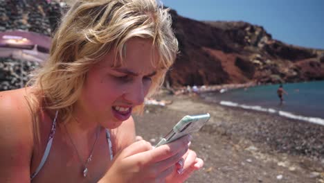 Mujer-Viajera-Sueca-Desliza-Su-Teléfono-En-La-Playa-Roja-En-Santorini-Grecia