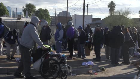 A-homeless-man-pushes-a-wheelchair-past-a-line-of-other-homeless-people,-Phoenix,-Arizona