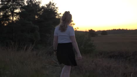 Young-blonde-woman-walking-throught-the-grass-in-the-nature-at-sunset-medium-shot