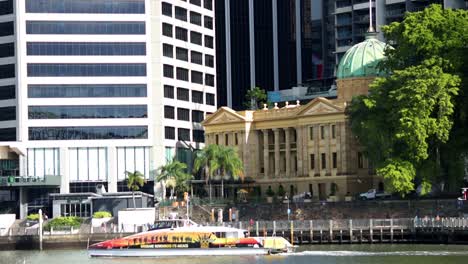 Altes-Zollhaus-In-Brisbane-–-Wunderschöne-Aufnahme-Des-Berühmten,-Von-Bäumen-Gesäumten-Gebäudes,-Während-Eine-Citycat-Fähre-Vorbeigleitet-Und-Das-Atemberaubende-Nebeneinander-Von-Alt-Und-Neu-Zeigt