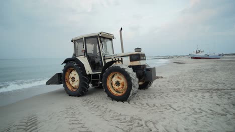 Rusty-tractor-standing-on-sand-at-a-beach-with-waves,-clouds-and-boats-in-the-background