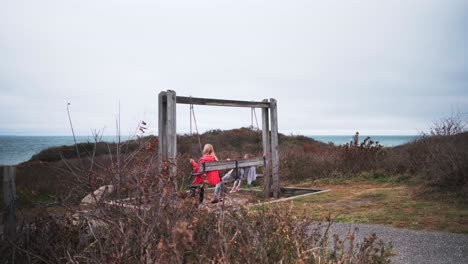 Eine-Frau-Mit-Zwei-Jungen-Mädchen-Auf-Einer-Holzschaukel-Mit-Blick-Auf-Das-Meer-Am-Montauk-Point