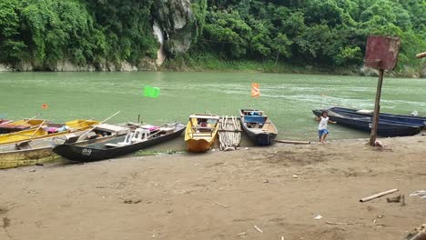 Niños-De-La-Aldea-Local-Corriendo,-Bailando-Cerca-Del-Río,-Barcos-Vistos-En-El-Fondo