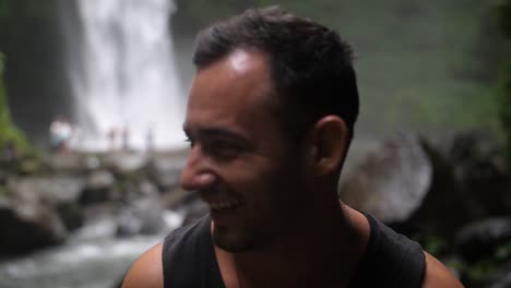 A-young-fit-man-is-laughing-and-smiling-towards-the-camera-as-he-stands-with-a-beautiful-and-big-waterfall-in-the-background