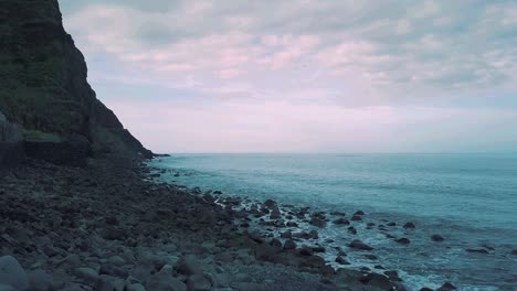 Volcanic-seascape-scene-at-sunrise-flying-forward-motion-past-stacked-stones-revealing-pebble-rocks-on-the-beach,-sky---clouds-of-Atlantic-ocean-Madeira-Island-landscape-HD-cine-grade-left-to-right