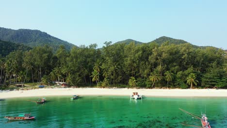 Fishing-and-touring-boats-anchoring-on-calm-beautiful-bay-with-green-water-washing-white-sand-of-tropical-island-with-palm-trees-in-Thailand