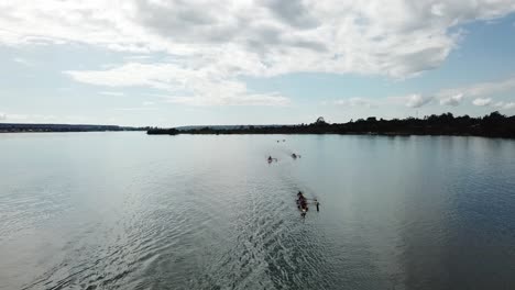 Aerial-view-over-group-of-long-rowing-boat-racing-teams-practising-on-lake-passing-below