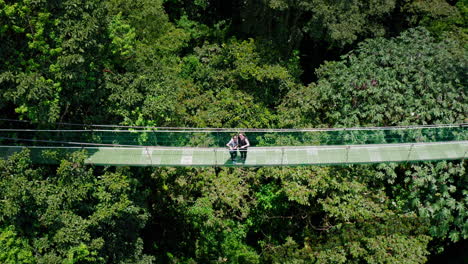 Toma-Aérea-De-Drones-Con-Cámara-De-Arriba-Hacia-Abajo-De-Una-Pareja-Disfrutando-De-La-Vista-En-La-Pasarela-Colgante-En-Medio-De-La-Selva-Tropical-En-Arenal,-Costa-Rica