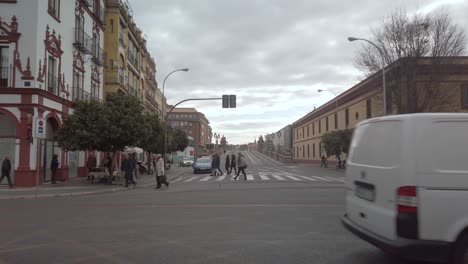 Cars-drive-by-as-pedestrians-cross-street-at-crosswak-in-background,-Seville