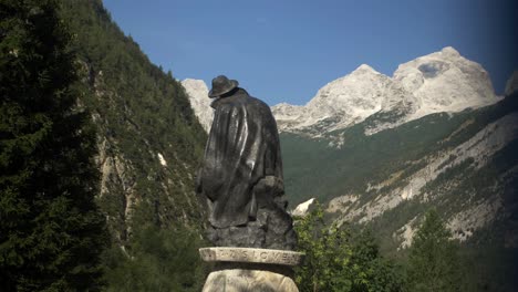 Statue-of-botanic-Julius-Kugy-revealed-from-behind-blue-flag,-looking-towards-mountains-on-sunny-summer-day