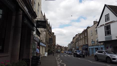 The-british-flag-is-hanging-on-top-of-a-hotel-in-the-English-village-Tetbury-with-a-few-people-standing-on-the-sidewalk-and-cars-passing-by-on-the-road-in-the-quiet-village-centre