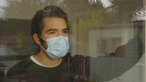 Male-looking-out-and-leaning-on-a-window-longingly-wearing-a-facemask-in-quarantine