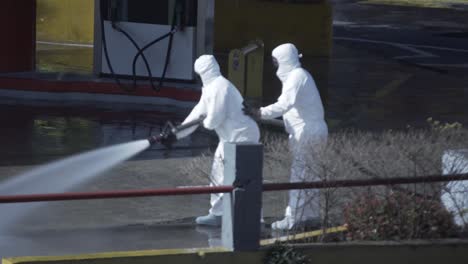 Zwei-Männer-In-Biohazard-Anzügen-Reinigen-Während-Der-Covid-19-Pandemie-Den-Boden-Einer-Tankstelle-Mit-Einem-Wasserstrahl,-Der-Aus-Einem-Feuerwehrschlauch-Kommt