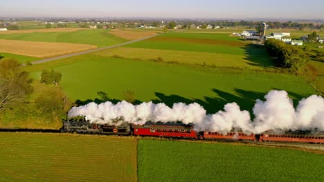 Luftaufnahme-Von-Ackerland-Bei-Sonnenaufgang-Mit-Einer-Dampfmaschine-Und-Einem-Personenzug,-Der-Mit-Voller-Dampf-Und-Rauch-Fährt