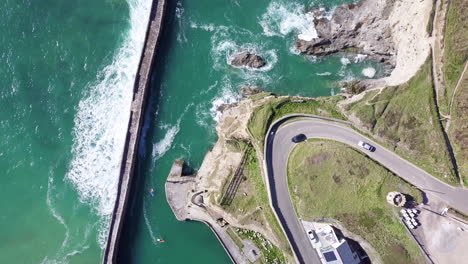 Cars-Driving-In-Lighthouse-Hill-By-Curve-Road-With-Ocean-Waves-On-Pier-Near-Village-Of-Portreath,-Cornwall,-England-In-Summer