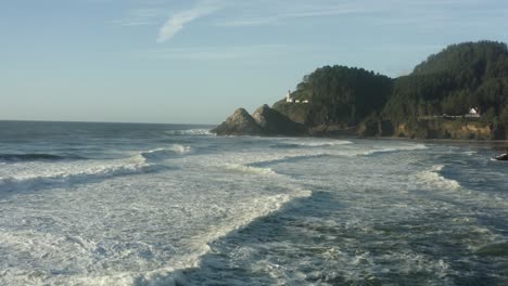Wide-aerial-moving-low-across-waves-toward-Haceta-Head-lighthouse-in-Oregon