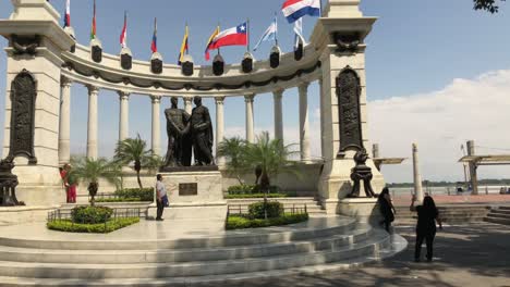 Toma-Panorámica-Del-Monumento-A-La-Rotonda-En-El-Malecón-Simón-Bolívar-De-Guayaquil-En-Un-Día-Soleado-Y-Las-Banderas-En-El-Fondo-Ondeando-Y-La-Gente-Tomando-Fotos