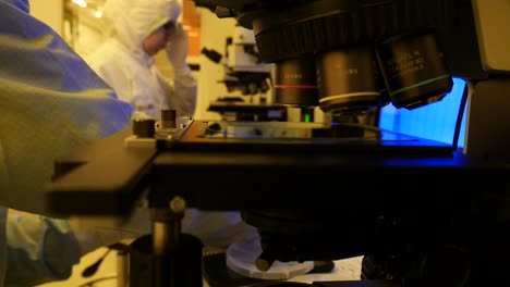 A-close-up-of-a-microchip-lying-under-a-microscope,-a-scientist-at-work-in-the-background