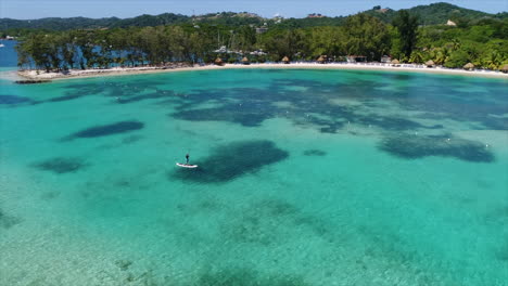 Vista-Aérea-En-Cámara-Lenta-De-Personas-Haciendo-Paddleboarding-Cerca-De-La-Playa-De-Un-Resort-En-El-Mar-Caribe-Hondureño