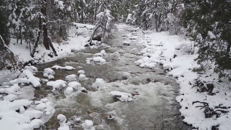 Slow-Motion-Pull-Back-shot-of-Wooded,-Freezing-Merced-River-In-Yosemite,-Rushing-through-Snow-covered-Rocks-and-Forest
