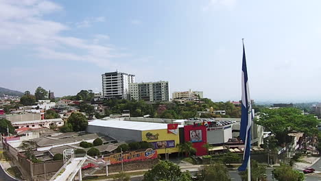 Toma-De-Drones-De-La-Bandera-Salvadoreña-Que-Sopla-En-El-Viento-Mientras-Mira-El-Horizonte-De-La-Ciudad