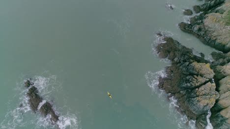 Top-down-aerial-descending-upon-a-person-in-a-yellow-kayak-sailing-around-a-rocky-UK-coastline-in-murky-blue-sea-waters