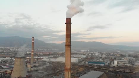 Rauchende-Industrieschornsteine,-Gebäude-Und-Berge-Als-Hintergrund---Luftaufnahme