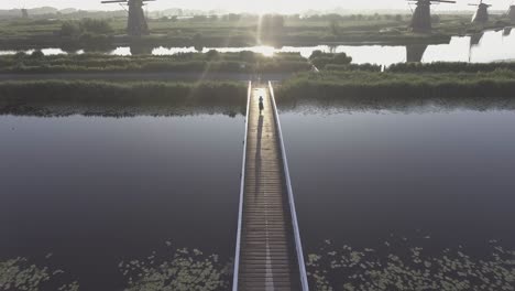 A-drone-shot-panning-down,-while-a-girl-walks-on-a-bridge,-with-Dutch-Windmills-in-the-Netherlands-during-sunrise