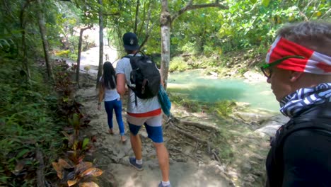 A-man-walking-across-a-hand-built-wooden-bridge-when-camera-pans-left-to-reveal-a-bright-blue-water-stream-flowing-underneath-through-the-lush-tropical-jungle-of-Bohol,-Philippines