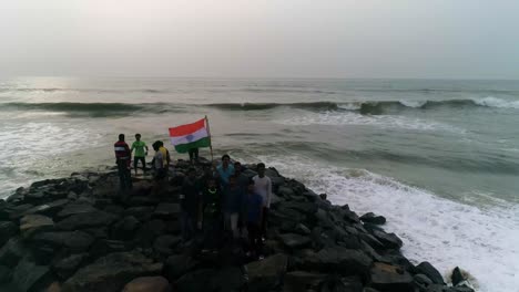 Grupo-De-Hombres-Jóvenes-Patriotas-Sostienen-La-Bandera-Nacional-India-De-Pie-En-Una-Playa-Rocosa-Con-Olas-Rompiendo