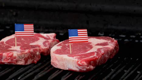 Two-juicy-rib-eye-steaks-sitting-on-the-grill-and-cooking-with-two-tiny-American-flags-tooth-picked-into-them