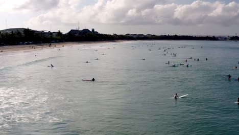 Many-Surfers-on-a-cloudy-day-waiting-for-large-waves-to-ride,-Aerial-drone-flyover-against-sun-shot