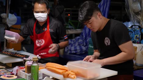 Two-chefs-preparing-fish-on-street-food-stall,-night-market-Bangkok