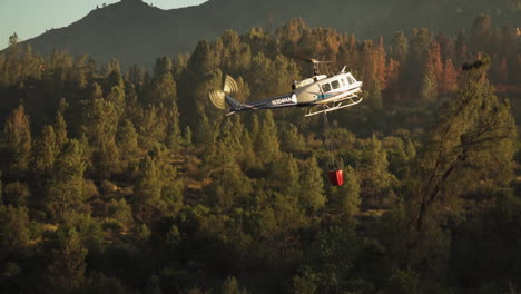 Helicopter-with-water-bucket-manoeuvring-over-forest-fire,-slow-motion
