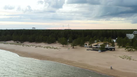 Vista-Aérea-De-Un-Restaurante-En-La-Playa-De-Yyteri,-Levantando-Y-Revelando-Molinos-De-Viento-Y-Un-Cielo-Colorido-Y-Nublado,-En-Pori,-Finlandia---Grúa,-Disparo-De-Drones