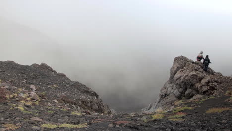 Dos-Personas-De-Pie-Al-Borde-De-Las-Rocas-En-El-Monte-Etna-Con-Vapor-Volcánico-En-El-Fondo
