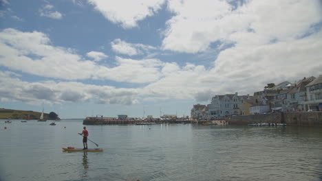 Man-paddleboards-across-water-near-St-Mawes-coastal-village,-wide-angle