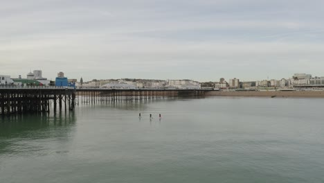 Gente-En-Paddle-Boards-Cerca-Del-Muelle-De-Brighton-Con-Gaviotas-Volando-Y-Playa-A-La-Vista