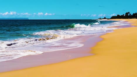 HD-Hawaii-Kauai-slow-motion-static-wide-shot-of-ocean-waves-crashing-and-washing-in-from-left-to-right-onto-beach-along-frame-right-with-an-island-and-a-few-clouds-in-distance