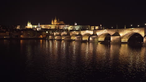 The-Prague-Castle-and-Charles-bridge-over-river-Vltava-in-the-historical-centre-of-Prague,-Czechia,-lit-by-lights-at-night,-shot-from-the-other-side-of-the-river,water-slowly-flowing-below-the-bridge