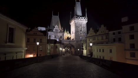 A-view-of-a-gothic-gate-tower-at-one-end-of-Charles-bridge-in-the-historical-city-centre-of-Prague-at-night-during-a-Covid-19-lockdown-with-no-people-anywhere,-picturesque-architecture,-tilt-4k-shot