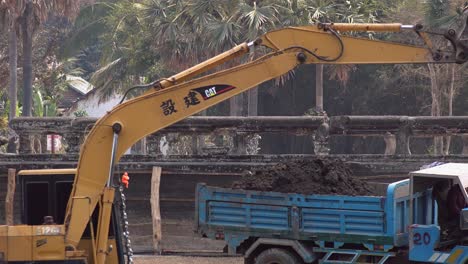 Medium-Shot-of-Digger-Emptying-its-Bucket-Full-of-Earth-Into-the-Bed-of-a-Blue-Truck-Then-Truck-Leaves