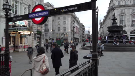 Una-Mujer-Con-Una-Mascarilla-Quirúrgica-Desciende-Con-Otra-Persona-A-Una-Estación-De-Metro-En-Un-Circo-Piccadilly-Vacío