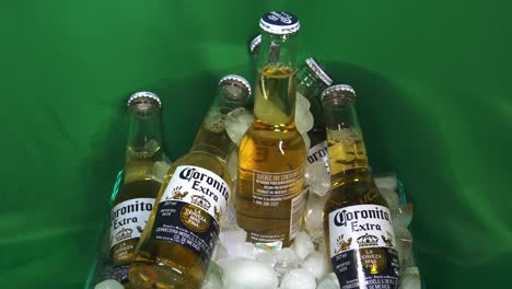 2-3-Refrescantes-6-Paquetes-De-Botellas-Pequeñas-De-Cerveza-De-Vidrio-Coronita-Extra-De-200-Ml-Que-Miran-Hacia-Un-Balde-Lleno-De-Hielo-Frente-A-Una-Pantalla-Verde-Con-Una-Botella-Central-Giratoria-Que-Muestra-La-Cerveza-Con-Tapa-De-Corona