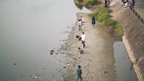 People-Skipping-Stones-By-The-Kamogawa-River-In-Kyoto,-Japan-During-Springtime---high-angle-slowmo-shot