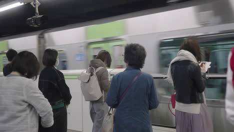 Standing-Commuters-On-The-Subway-Station-Platform-With-Traveling-Train-Passing-By-In-Tokyo,-Japan