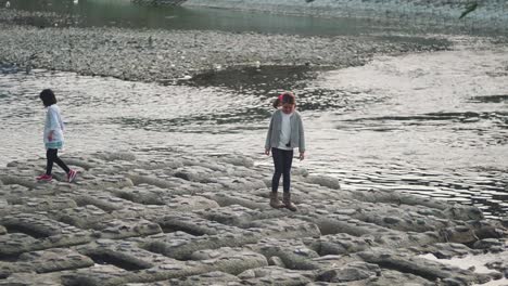 Two-Kids-Having-Fun-On-The-Rocky-Surface-By-The-Kamogawa-River-In-Kyoto,-Japan---wide-slowmo-shot