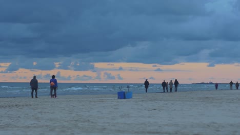 People-walking-on-the-beach,-two-people-doing-Nordic-walking,-colorful-autumn-sunset,-wide-shot